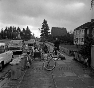 Gerhart-Hauptmann-Straße: Sperrmüll: Kinder beim Durchwühlen der Haufen: hinten links Straßeneinmündung: rechts Jägerzaun, Vorgarten, Häuser: Fahrradweg, Straßenlampen, Fahrräder, Mülltonnen, parkende Autos, Fußgänger