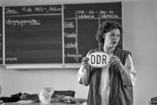 Geschichtsunterricht an einer Realschule. Die Lehrerin hält ein DDR-Hoheitszeichen für PKW hoch