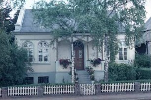 Lübecker Straße 39: traufständiges Wohnhaus: Straßenfront mit Rundbogenfenstern, Eingangsveranda, Treppe und Eingangstür: davor Vorgarten, zwei Birkenbäume, Lattenzaun