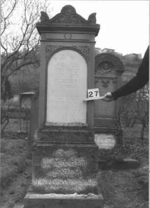 Grabstein 27 (Index: 99805080) Material: Sandstein mit weißer Marmorplatte Zustand: Sockel verwittert mit Textverlust Maße HxBxT: 200x63x25 cm Beschreibung: Stele auf gestuftem Sockel mit Aufsatz, Inschriftennische mit Rundbogen Symbolik: Blätterornamentik beidseitig der Inschriftentafel, Voluten und Akroterien als Stelenaufsatz: Grabstein 27