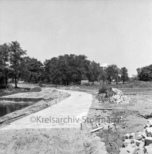 Wanderweg Glinder Au: Ausbau mit Steinen: rechts Erd- und Sandhaufen sowie Baumaterial: links Regenrückhaltebecken: hinten Bäume: rechts dahinter Wohnblock