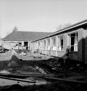 Volksschule am Schweinemarkt [später Emil-Nolde-Schule]: Erweiterungsbau: vor Fertigstellung: Außenansicht: davor hügelige Wiese, Zaun: im Hintergrund rechts Turnhalle
