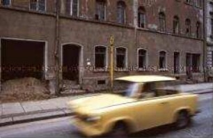Last Exit DDR 1990 - gelber Trabant fährt auf der Straße vor einem leerstehenden Haus
