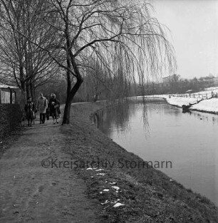 Trave: rechts Travewanderweg mit Personen, Bäumen, Zaun und Schuppen: hinten rechts Wiese: dahinter Gewerbegebäude: Winter
