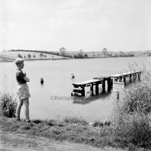 Moorteich mit Schulschwimmsteg: im Vordergrund links Junge am Ufer: rechts Schilf: hinten Knicklandschaft