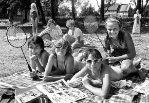 Sommer : Gruppenbild von vier Jugendlichen die in der Sonne liegen. Sie haben Badesachen an, Federballschläger in der Hand und haben Sonnenbrillen auf.