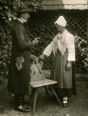 Ehepaar stehend vor historischem Holzstuhl, Frau mit Haube rechts gestützt, Mann mit Pfeife links daneben. Halbtotale