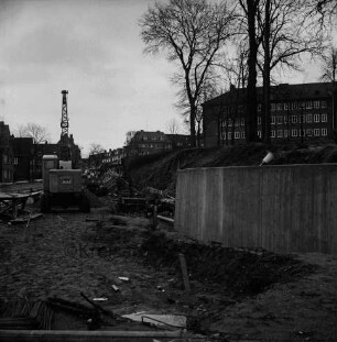 Mewesstraße: Stützmauer beim Kreishaus: Blick Richtung Reimer-Hansen-Straße: vorn Baustelle mit Baumaterial, Holzstützen und Bagger: rechts Kurparkhügel mit Bäumen: dahinter Kreishaus: hinten links Wohnhäuser