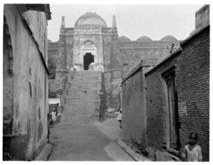 Delhi, Indien. Jama Masjid (Masjid-i Jahan Numa, "Freitagsmoschee") (1644-1658; Shah Jahan). Blick durch eine Gasse mit Wohnhäusern auf einen Zugang zur Freitagsmoschee in massiver Festungsmauer mit Kuppel und Treppenstufen