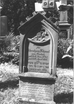 Grabstein 94 (Index: 99787177) Material: Sandstein und aufgesetzter Zapfen aus Metall Zustand: stärker verwittert am Sockel Maße HxBxT: 142x57x18 cm Besonderheit: Schrift schwarz: Grabstein 94