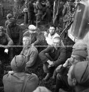 Auf dem Boden sitzende Kriegsgefangene neben einem LKW der Wehrmacht