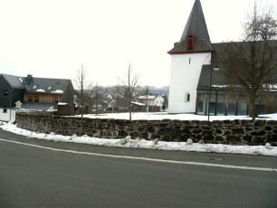 Ansicht von Norden mit Kirche (Kirchturm im Kern Frühgotisch - Langhaus 1970 neu erbaut) über Kirchhofmauer (Wehrgang mit Steinbrüstung im 18 Jh abgetragen) im Verlauf nach Osten