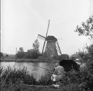 Holländerwindmühle. Ansicht über einen Fluß. Im Vordergrund ein Angler