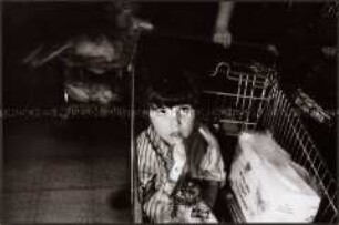 Kleinkind mit einer Packung Strohhalmen und Windeln im Einkaufswagen eines Supermarktes (Sonderthema: Essen und Trinken)