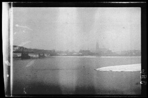 Fluss mit Stahlbrücke und Betonsockeln mit zweitürmiger Kirche im Hintergrund