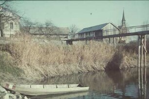 Deutschland. Ruderboot unterhalb einer hölzernen Brücke an einem Ufer vor einem Gehöft. Blick zur Dorfkirche