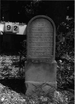 Grabstein 92 (Index: 100086572) Material: Sandstein Zustand: Sockel abgeblättert Maße HxBxT: 98x38x11 cm Beschreibung: Stele, getreppter Sockel, Rundbogenabschluß Steinmetz: FB ?? Besonderheit: Kindergrab; - steht im 90ø Winkel zu den anderen Steinen: Grabstein 92