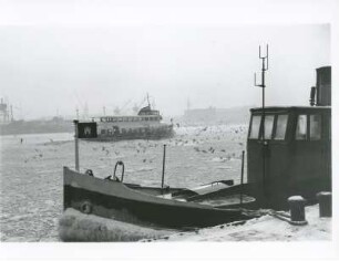 Hamburg. Der Hafen der Freien- und Hansestadt im Winter. Eine Hafenfähre bahnt sich ihren Weg durch das vereiste Hafenbecken. Im Vordergrund ein vertäuter Schlepper. Möwen suchen nach Essbarem