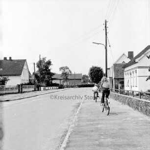 Dorfring: rechts Mädchen und Junge auf Fahrrädern auf Fußgängerweg: dahinter Strommast: am Straßenrand Bauernhäuser
