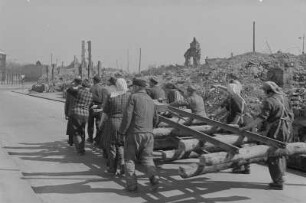 Berlin. Aufbauhelfer transportieren Bahngleise mit Holzschwellen auf einer Straße in Trümmerlandschaft